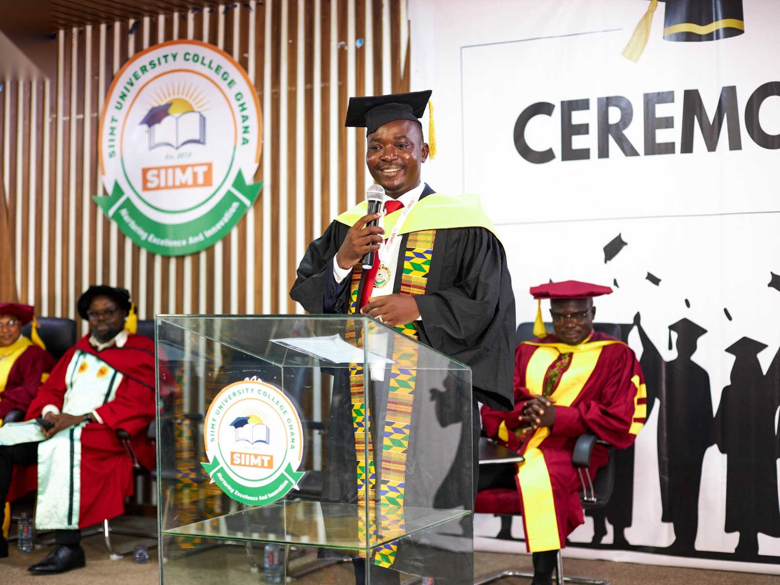 A graduate in a gown and kente stole speaking at the podium at the SIIMT Graduation 2024